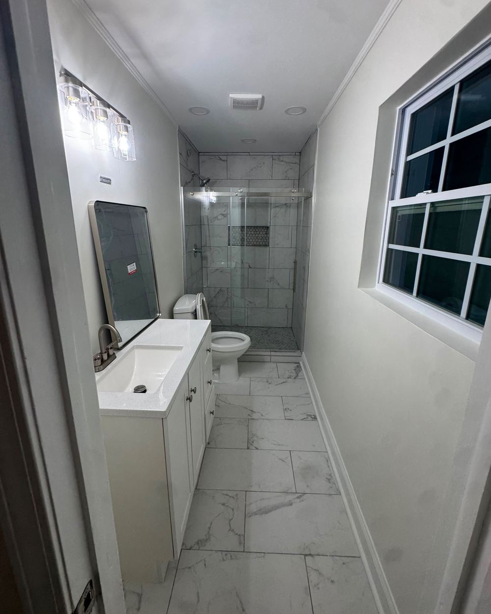 Hall bathroom after renovation: white vanity, new light fixture, glass shower, and marble-look tile floor