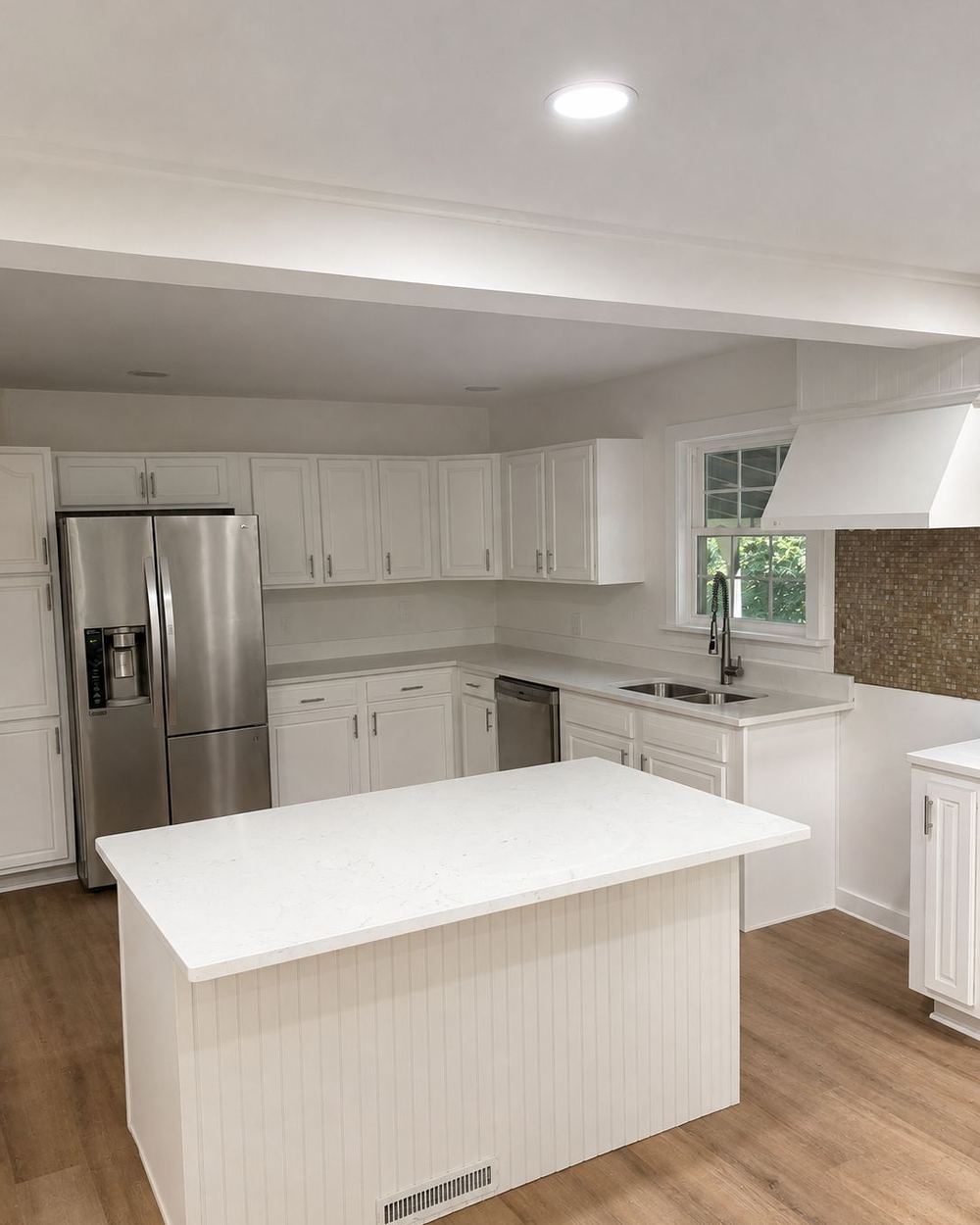 Kitchen after renovation: white cabinets and countertops, a new white island, and wood-look floors