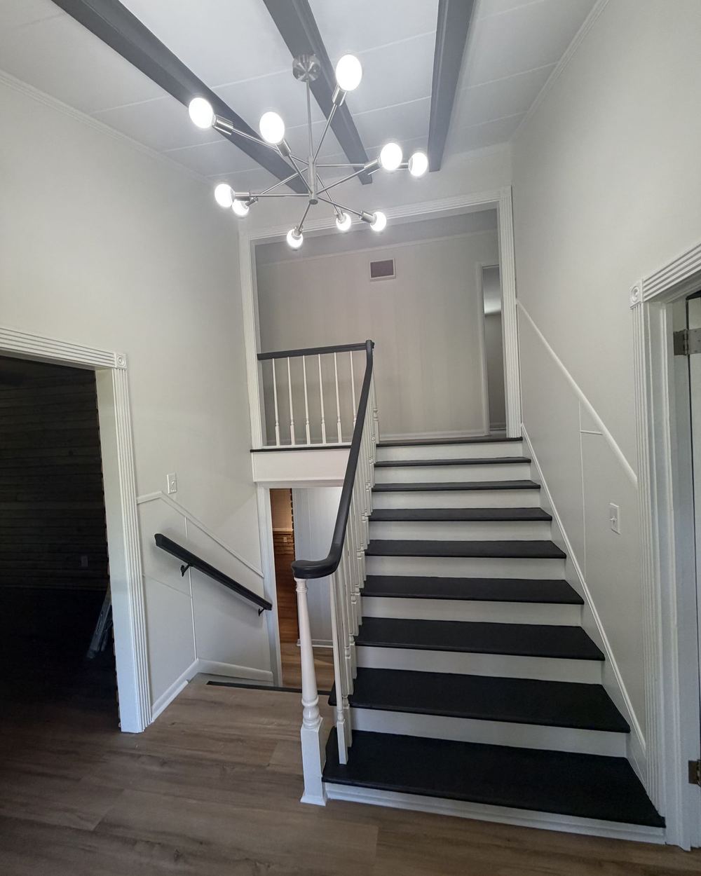 Foyer staircase after renovation: black treads with white risers, white railings, painted beams, and a modern chandelier