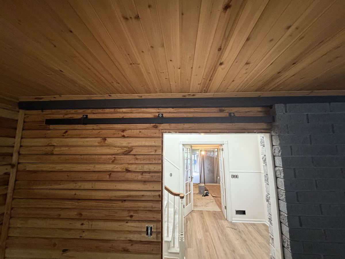 Den with warm wood paneling on the walls and ceiling, opening to a bright hallway